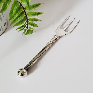 Puede incluir: Un tenedor de servir plateado con tres púas y un mango decorativo. El mango presenta una serie de líneas verticales y un extremo redondeado. El tenedor está colocado sobre una superficie blanca, con una planta en maceta en el fondo.