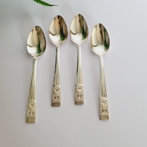 Puede incluir: Cuatro cucharas plateadas con diseños florales ornamentados en los mangos, dispuestas sobre una superficie gris claro. Las cucharas son reflectantes, y los mangos tienen forma rectangular con grabados detallados. Follaje verde visible en la esquina superior izquierda.