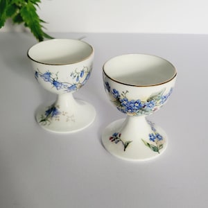 Puede incluir: Dos hueveras de porcelana blanca con un diseño floral con flores azules y ribete dorado. Las hueveras tienen una base de pedestal y están decoradas con delicados estampados florales azules. Las hueveras son de estilo vintage.