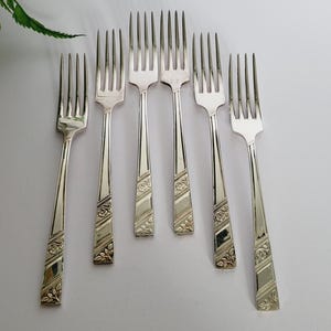 Puede incluir: Siete tenedores plateados dispuestos sobre una superficie blanca. Cada tenedor presenta un diseño floral decorativo en el mango. Las púas están espaciadas uniformemente. Los tenedores están limpios y pulidos, midiendo unos 20 cm de largo.