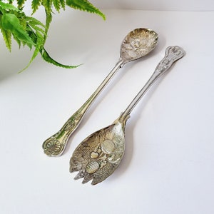 Puede incluir: Dos utensilios de servir antiguos de plata. Uno es una cuchara con un diseño floral detallado en el cuenco. El otro es un tenedor de servir con un patrón floral similar. Ambos tienen mangos ornamentados y están sobre un fondo blanco.