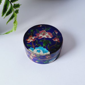 May include: A small, round, blue trinket box with a floral design. The lid features pink and white flowers with green leaves against a dark blue background. The box has a gold-coloured rim and is set on a white surface.