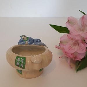 May include: A ceramic mushroom-shaped dish with a sleeping figure in blue and green. The dish has a beige exterior with green and blue window details. Pink flowers are to the right.