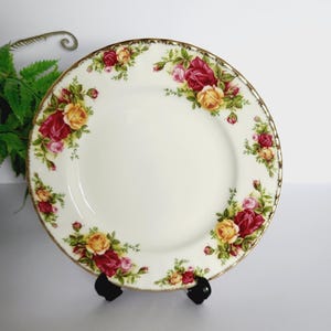 以下が含まれることがあります： 赤、黄色、ピンクのバラのフローラルパターンが施された白いセラミックプレート。 プレートは金色の縁取りがあり、黒いスタンドに展示されています。 花柄はプレートの縁に沿って配置されています。