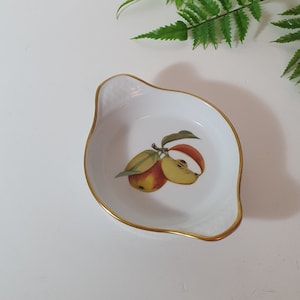 May include: White ceramic dish with a gold rim, featuring a painted illustration of apples. The dish has a leaf-like shape with a small handle. A sprig of green fern is in the background.