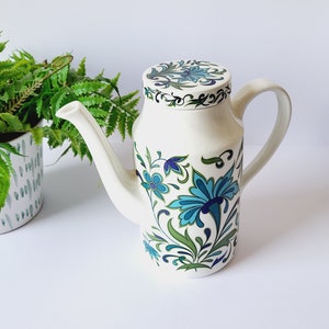 May include: A white ceramic teapot featuring a blue and green floral pattern. The teapot has a long spout, a curved handle, and a matching lid. The floral design is repeated on the lid. The teapot is a decorative item, suitable for serving tea.