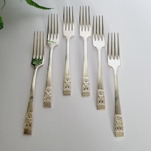Peut inclure: Six fourchettes argentées avec des motifs floraux ornent les manches, disposées sur une surface blanche. Ces couverts, probablement pour un repas formel, sont présentés avec une fougère en arrière-plan.