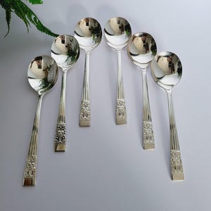 Puede incluir: Seis cucharas soperas plateadas dispuestas sobre una superficie blanca. Las cucharas tienen un cuenco redondeado y un mango decorativo con un diseño floral. Las cucharas reflejan el entorno.