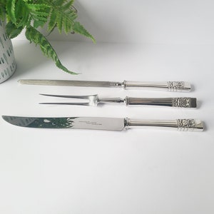 May include: A set of three silver serving utensils: a carving knife, a carving fork, and a sharpening steel. The handles have a decorative floral design. The knife blade has text. The items are arranged on a white surface.