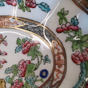 May include: Close-up of a white ceramic dish with a floral design. The dish features pink flowers, green leaves, and blue accents. A gold border with black and white detailing frames the floral pattern. The dish has a brown rim.