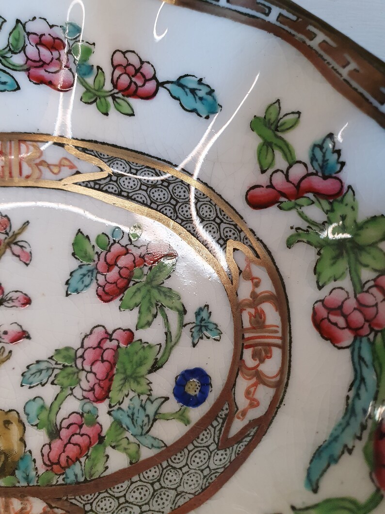 May include: Close-up of a white ceramic dish with a floral design. The dish features pink flowers, green leaves, and blue accents. A gold border with black and white detailing frames the floral pattern. The dish has a brown rim.