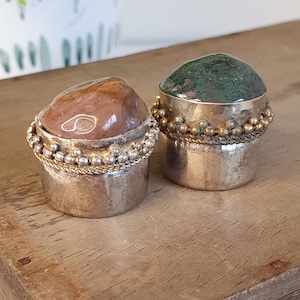 May include: Two decorative stone bottle stoppers. One has a reddish-brown stone top, the other a green stone top. Both have silver-coloured metal bases with gold-coloured beaded accents. The stoppers are displayed on a wooden surface.