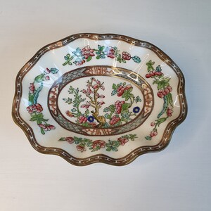 May include: A decorative, scalloped-edge dish with a floral design. The dish features a central oval with a tree and floral motif, surrounded by a border of flowers and foliage. The rim has a brown and gold trim. The dish is white with pink, blue, and green accents.