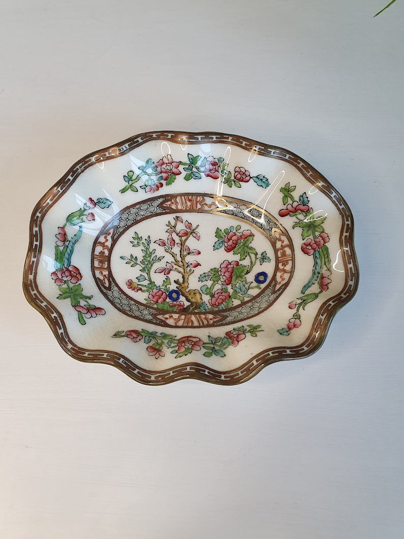 May include: A decorative, scalloped-edge dish with a floral design. The dish features a central oval with a tree and floral motif, surrounded by a border of flowers and foliage. The rim has a brown and gold trim. The dish is white with pink, blue, and green accents.