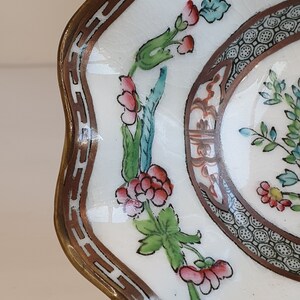 May include: A white ceramic dish with a scalloped edge and a brown border. The dish features a floral design with red, pink, and green accents. The rim has a decorative brown pattern. The dish is likely vintage or antique.
