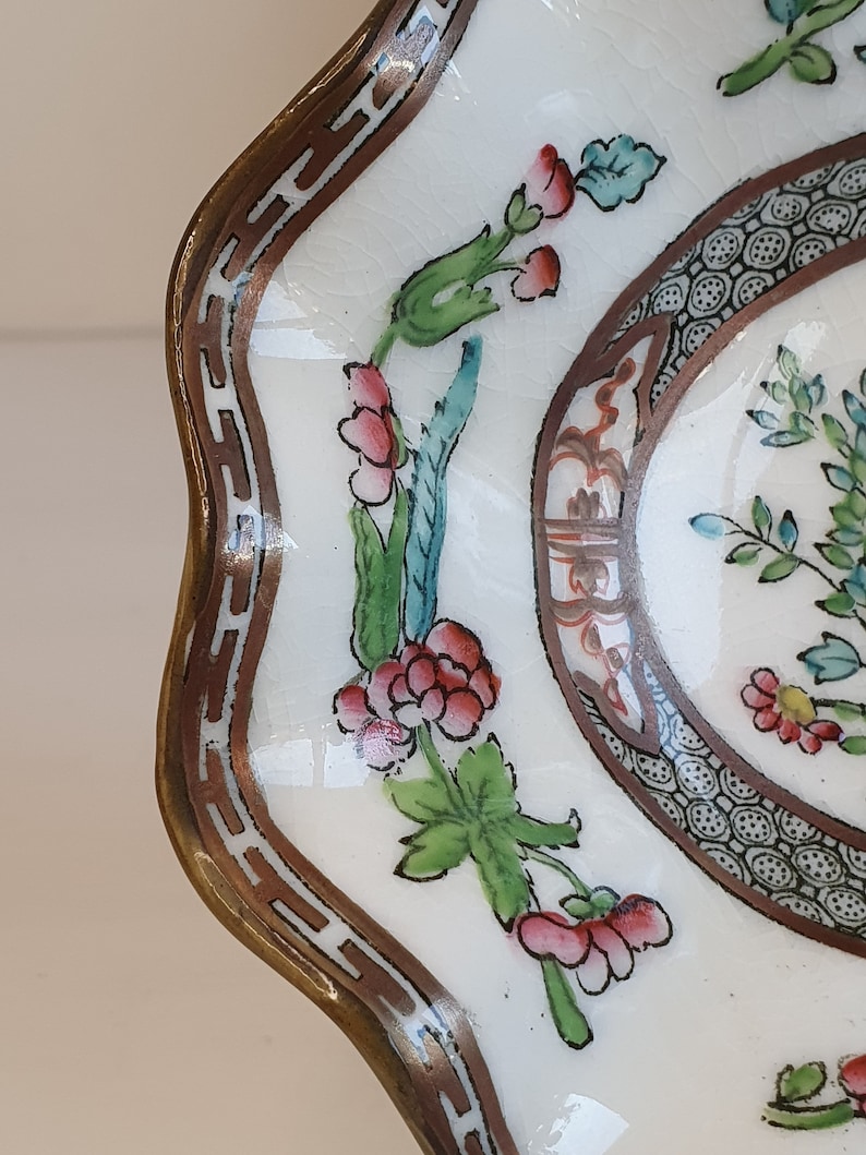 May include: A white ceramic dish with a scalloped edge and a brown border. The dish features a floral design with red, pink, and green accents. The rim has a decorative brown pattern. The dish is likely vintage or antique.