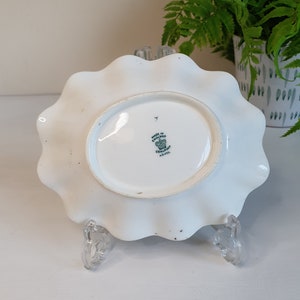 May include: White, scalloped-edge ceramic plate with an oval center. The underside features a stamp that reads "Made in England" and "Coalport." The plate is supported by a clear, decorative stand.