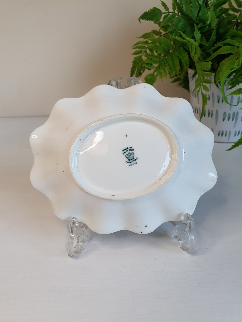 May include: White, scalloped-edge ceramic plate with an oval center. The underside features a stamp that reads "Made in England" and "Coalport." The plate is supported by a clear, decorative stand.
