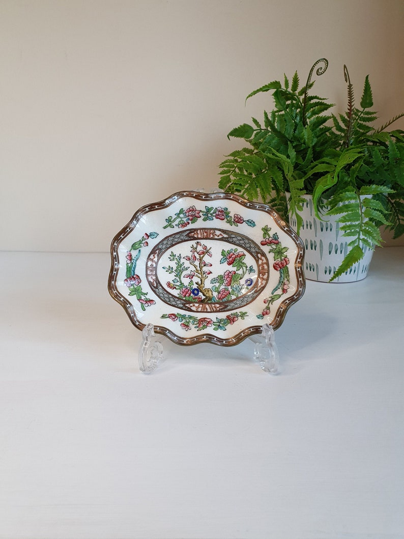 May include: An oval-shaped decorative dish with a scalloped edge. The dish features a central floral design with pink, blue, and green accents, framed by a brown border. The dish is displayed on a clear plastic stand, with a green plant in the background.