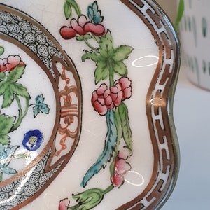 May include: A close-up of a white, scalloped-edge dish with a floral design. The dish features a brown and gold border and is decorated with pink, red, blue, and green flowers and leaves. The dish has a crackled surface.