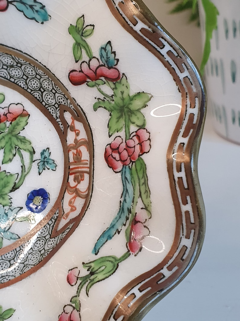May include: A close-up of a white, scalloped-edge dish with a floral design. The dish features a brown and gold border and is decorated with pink, red, blue, and green flowers and leaves. The dish has a crackled surface.