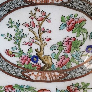 May include: Close-up of a white oval plate with a floral design. The plate features pink, red, and blue flowers, green leaves, and a brown border with a decorative pattern. The center has a tree with pink leaves.