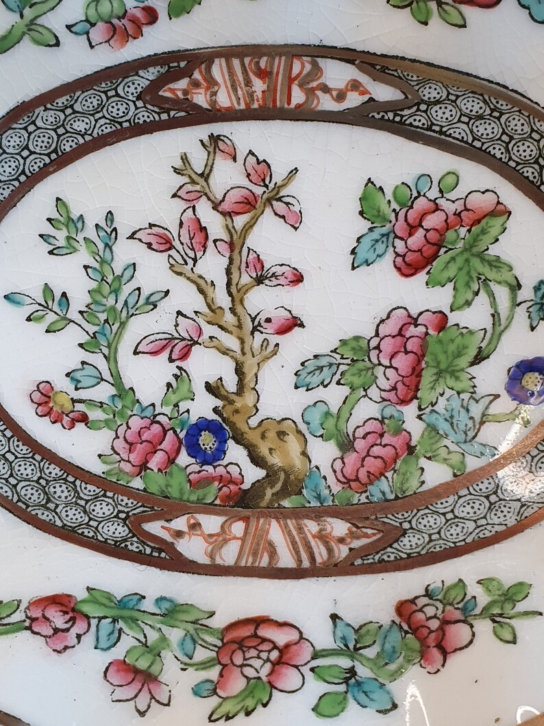 May include: Close-up of a white oval plate with a floral design. The plate features pink, red, and blue flowers, green leaves, and a brown border with a decorative pattern. The center has a tree with pink leaves.