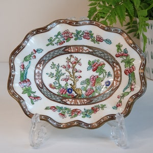 May include: A small, antique, white porcelain dish with a scalloped edge and a decorative floral design. The dish features a central oval with a tree and flower motif, surrounded by a brown border and pink and green floral accents. It is displayed on a clear plastic stand.