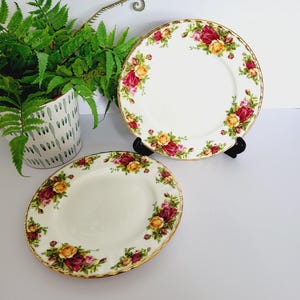 以下が含まれることがあります： 赤と黄色のバラのフローラルデザインが施された、金色の縁取りのある2つの白いセラミックプレート。白い表面に配置され、背景には鉢植えのシダ植物があります。