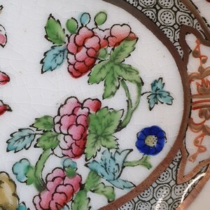 May include: Close-up of a white ceramic plate with a floral design. The plate features pink and red flowers with green leaves and blue accents. A brown border with a geometric pattern frames the design.