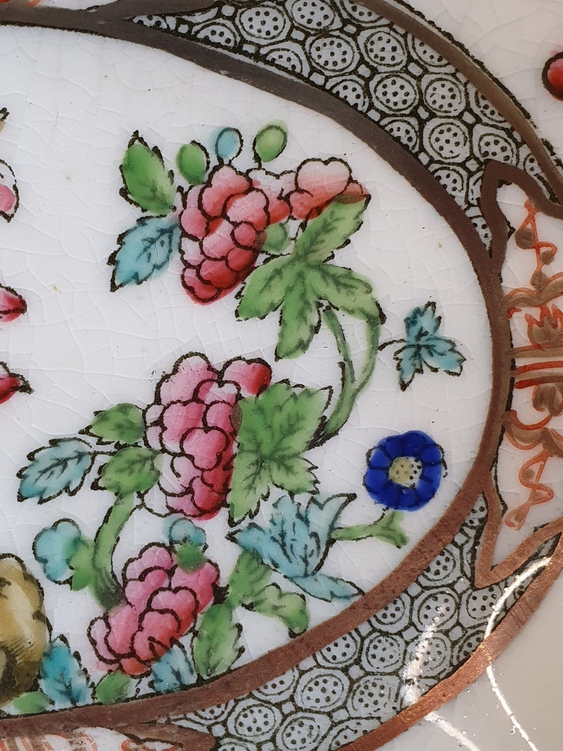 May include: Close-up of a white ceramic plate with a floral design. The plate features pink and red flowers with green leaves and blue accents. A brown border with a geometric pattern frames the design.