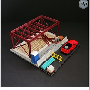 May include: A miniature construction site diorama featuring a red steel frame structure, a red pickup truck, a blue portable toilet, white concrete barriers, a yellow safety fence, stacks of white blocks, blue pipes, and gray metal barrels.