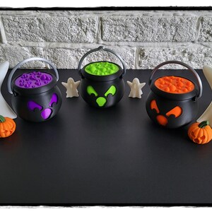 May include: Three black plastic cauldrons with silver handles, each featuring a different colored lid and face: purple, green, and orange. Two white ghost figurines, two small orange pumpkins, and two tiny white ghosts accompany them.
