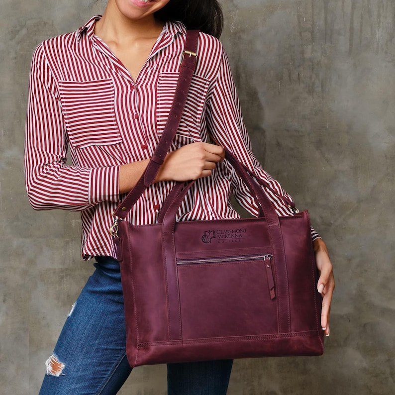 May include: A burgundy leather tote bag with a shoulder strap and a front zippered pocket. The bag has a rectangular shape and a logo on the front. The person is wearing a red and white striped shirt and blue jeans.
