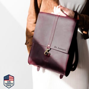 May include: A burgundy leather backpack with a flap closure and gold-colored hardware. The backpack has a top handle and adjustable straps. The text "Dr. Lennert" is visible on the front. The bag is held by a person.