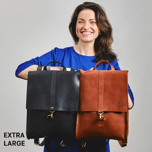 A woman in a blue top is smiling and holding two large leather backpacks, one black and one brown.