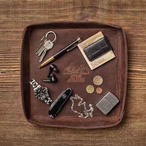 May include: A brown leather tray with a variety of items, including keys, a pen, a money clip with a 50 Euro bill, a watch, a Swiss Army knife, a chain, a lighter, and a pair of wireless earbuds. The tray has the words "Let's Go Adventure" embossed on it.