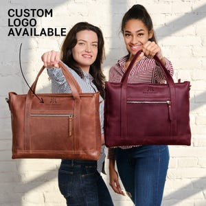 Two women are holding up two different types of leather handbags, one brown and one burgundy, against a white brick wall.