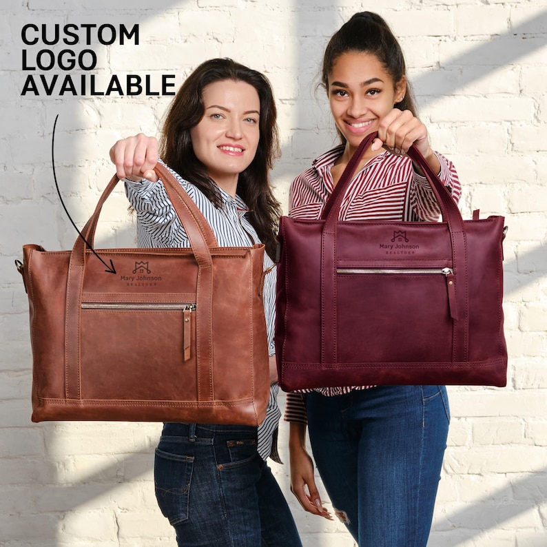 Two women are holding up two different types of leather handbags, one brown and one burgundy, against a white brick wall.