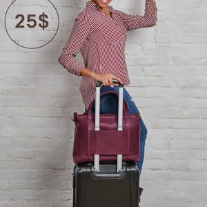 A woman in a striped shirt stands next to a black suitcase, smiling and holding her hair up.