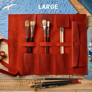 Peut inclure: Un &eacute;tui enroulable en cuir rouge avec plusieurs fentes pour ranger des pinceaux. L'&eacute;tui est enroul&eacute; et poss&egrave;de un rabat avec une fermeture &agrave; bouton. L'&eacute;tui est pos&eacute; sur une surface en bois avec des pinceaux &eacute;parpill&eacute;s autour.