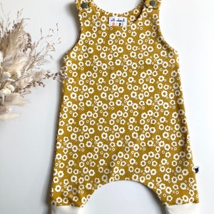 May include: Mustard yellow baby overalls with a white floral print and white ribbed cuffs. The overalls have a small label that says "all about pepi".