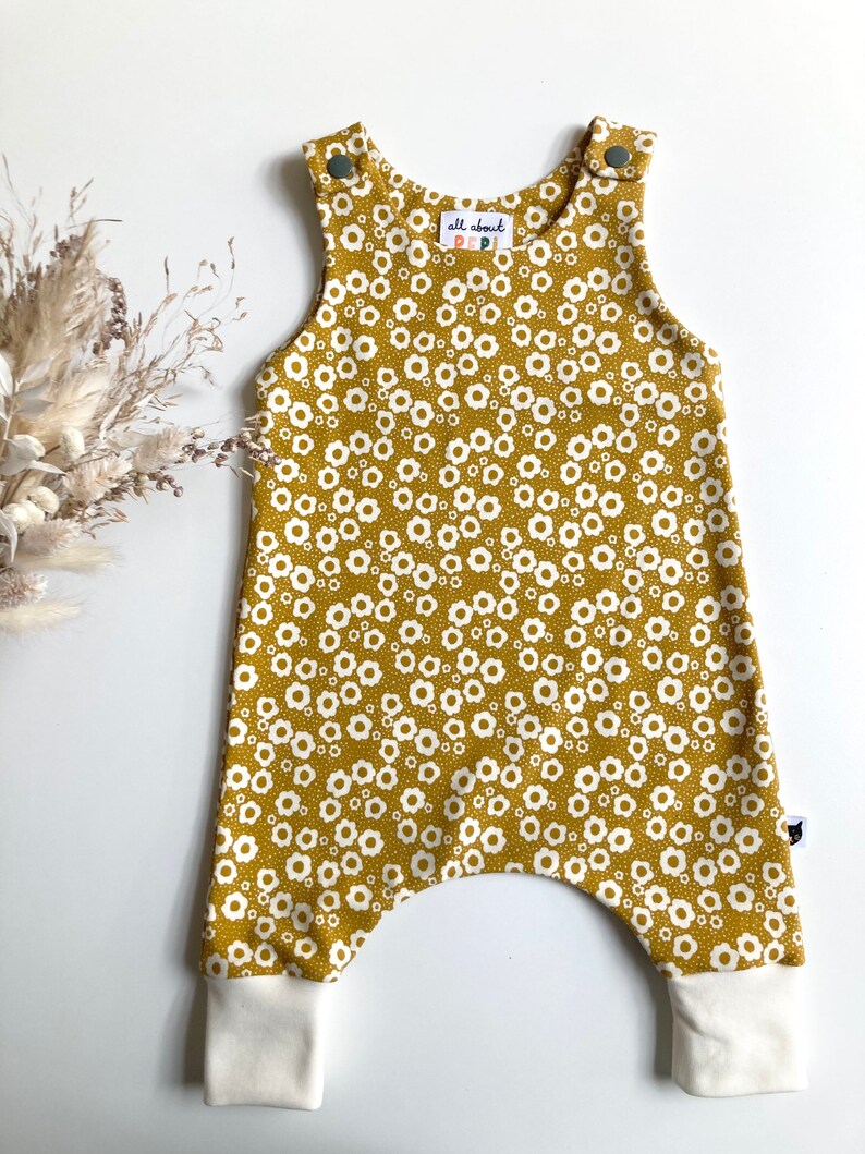 May include: Mustard yellow baby overalls with a white floral print and white ribbed cuffs. The overalls have a small label that says "all about pepi".