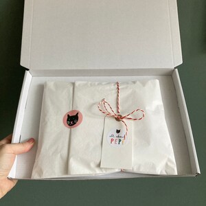 May include: A white cardboard box with the lid open, revealing a white tissue paper package tied with red and white string. The package has a pink circle sticker with a black cat face and a white tag with the text "all about PEPI!" in colorful letters.