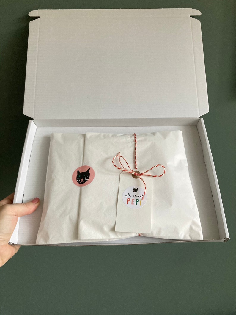May include: A white cardboard box with the lid open, revealing a white tissue paper package tied with red and white string. The package has a pink circle sticker with a black cat face and a white tag with the text "all about PEPI!" in colorful letters.