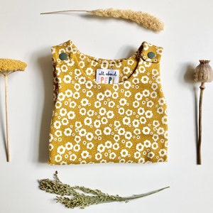 May include: A mustard yellow baby overall with a white daisy print. The overall has two snap buttons on the straps and a label that says "all about PIPI".