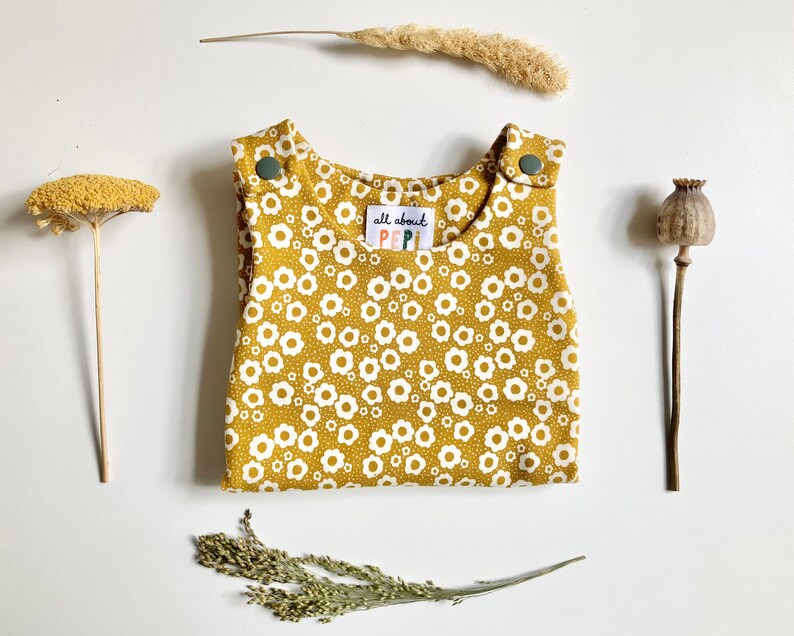 May include: A mustard yellow baby overall with a white daisy print. The overall has two snap buttons on the straps and a label that says "all about PIPI".