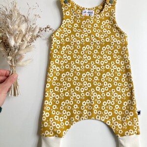 May include: Mustard yellow baby overalls with a white floral print and white ribbed cuffs. The overalls have a small label that says "all about baby".