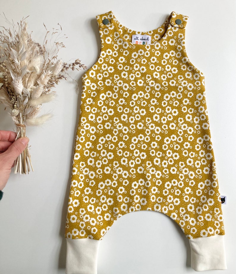 May include: Mustard yellow baby overalls with a white floral print and white ribbed cuffs. The overalls have a small label that says "all about baby".