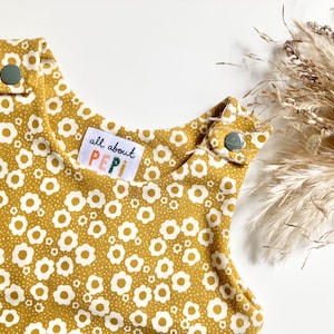 May include: A mustard yellow baby onesie with a white floral pattern. The onesie has a label that says "all about PEPI".